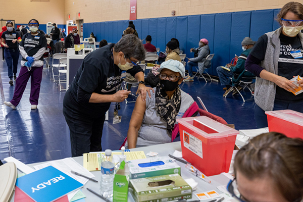 A person receives a vaccination at a Penn Medicine vaccine site by a masked professional while other masked people wait on folding chairs in the room.