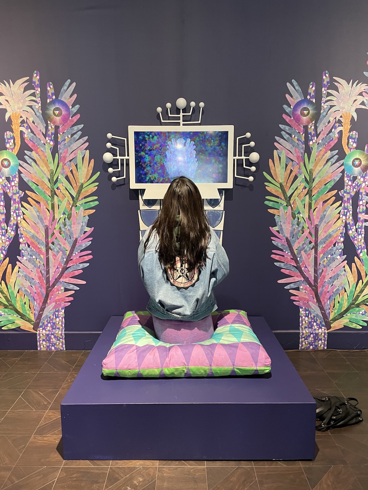 Woman sits on a cushion in front of a video screen flanked on both sides by and art installation of fantastical trees with yellow. pink, purple leaves