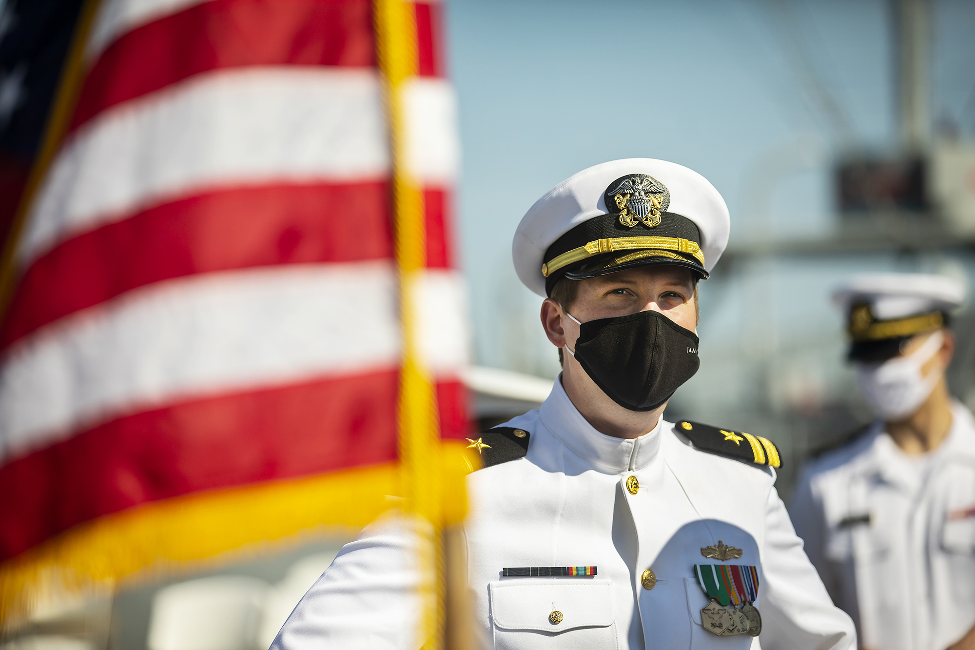 Person in uniform and face mask aboard a ship next to an American flag.