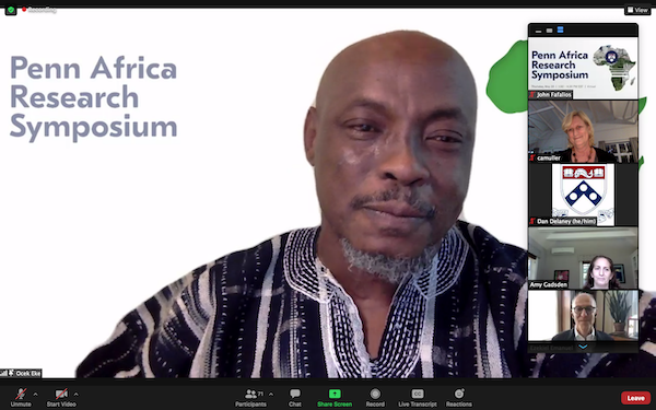 A Zoom video event shows man in a traditional African print shirt with the words "Penn Africa Research Symposium" on the background behind him, as six participants are seen in video images on the right side of the screen, stacked vertically.