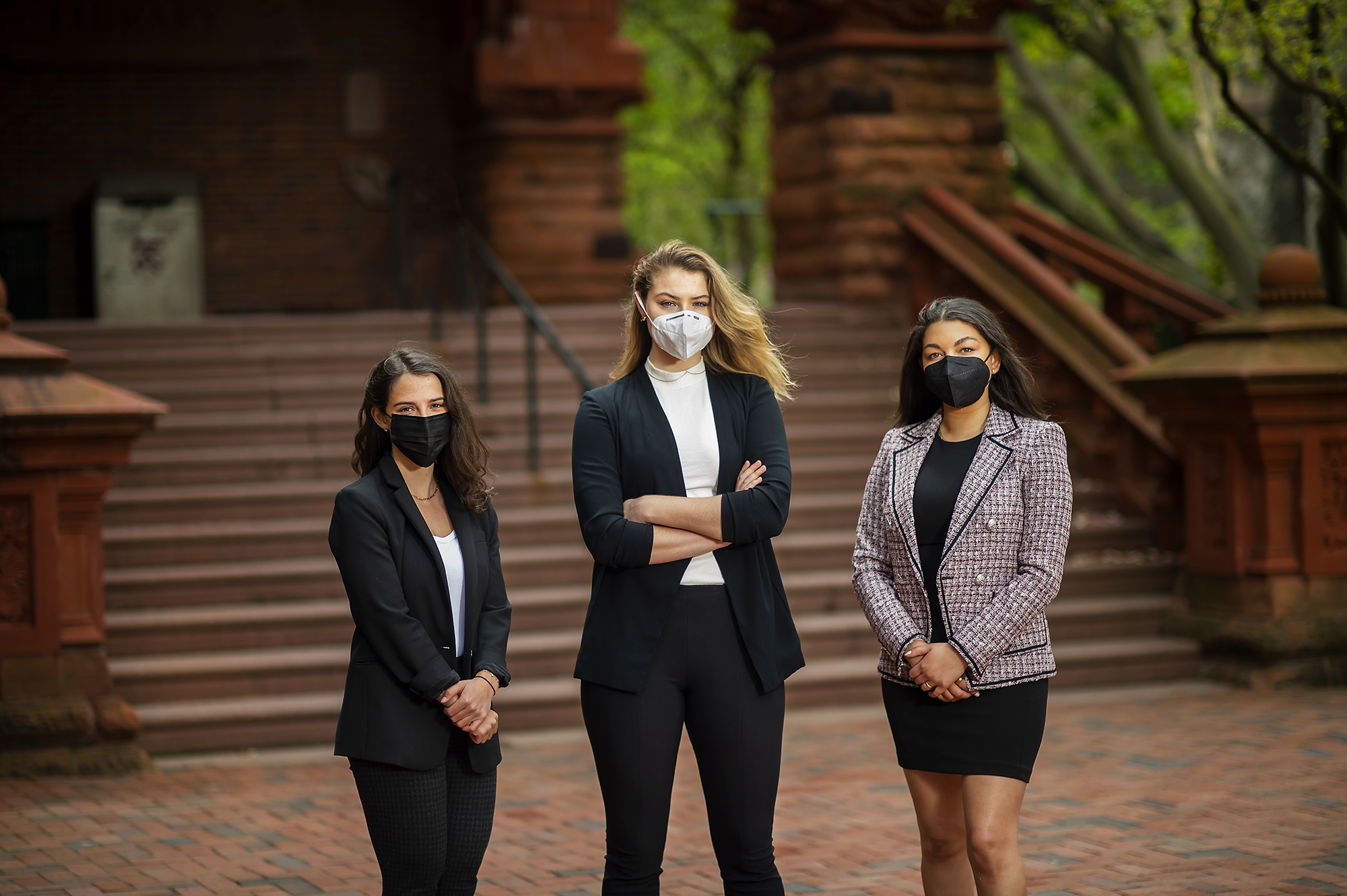 Three women wearing face masks and office dress clothes stand on a brick pathway with a wide brown staircase behind them