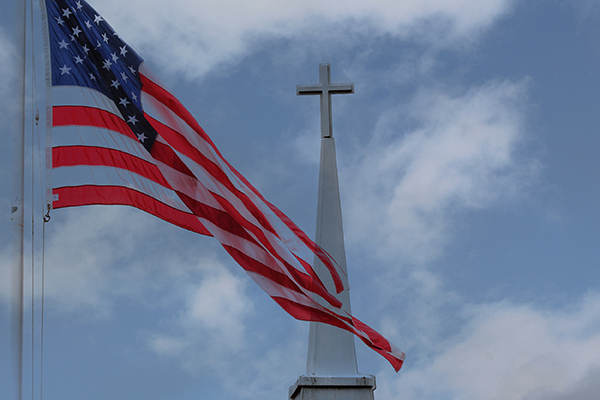 An American flag flaps in front of the tip of a white church steeple topped by a cross, with a blue sky and clouds in the background