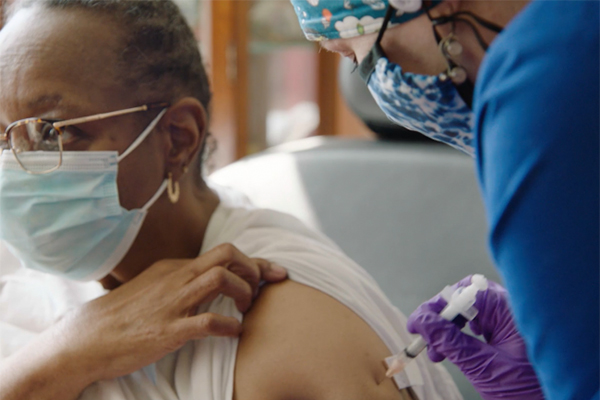 A masked health care worker give a vaccine to a person in the upper arm who is wearing a face mask.