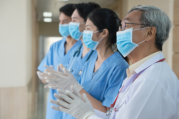 Three masked nurses in scrubs and one masked doctor standing in a line smiling and clapping.