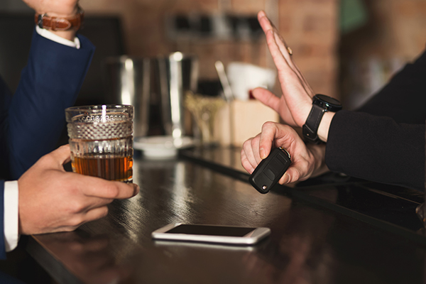 Person at bar waving away a drink offered while holding car keys with smartphone laying on bar next to them.