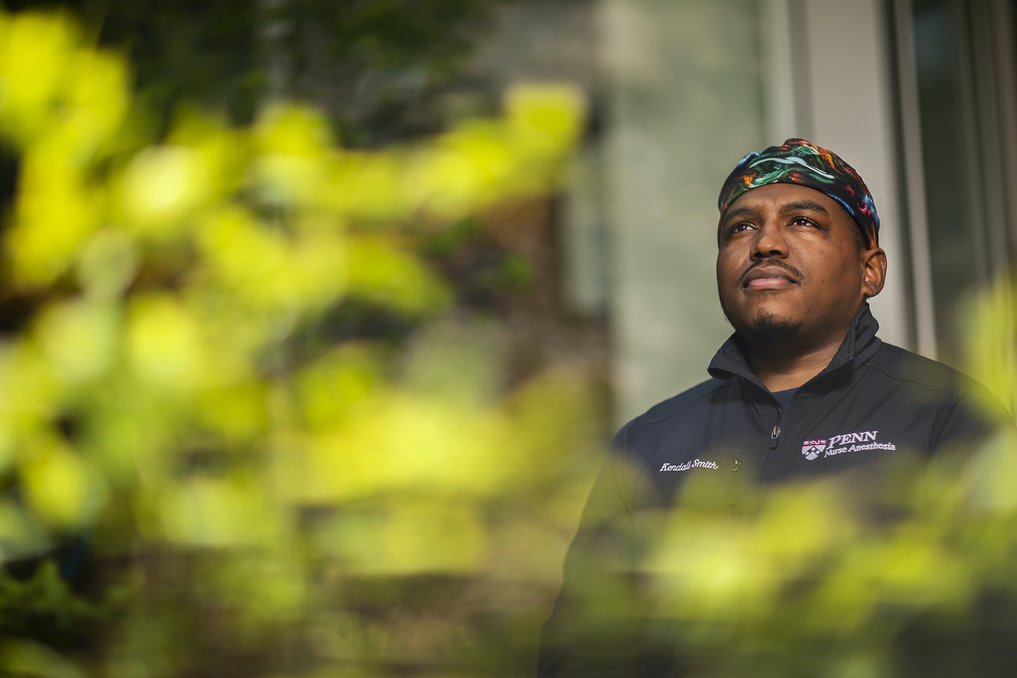 A person standing outside, with blurred leaves in the foreground. The person is wearing a surgical cap and the jacket reads, "Kendall Smith, PENN Nurse Anesthesia"