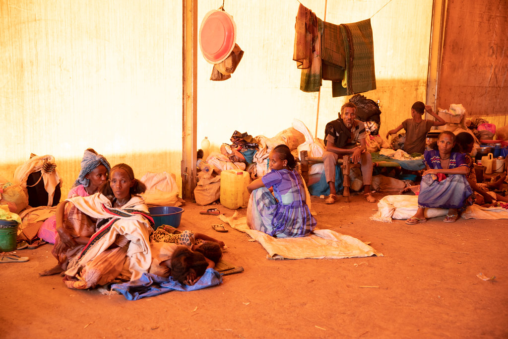 Ethiopian refugees, mostly women and children from the Tigray region, sit and lay on blankets on a dirt floor, as some blankets and clothing hang on a clothesline behind them.