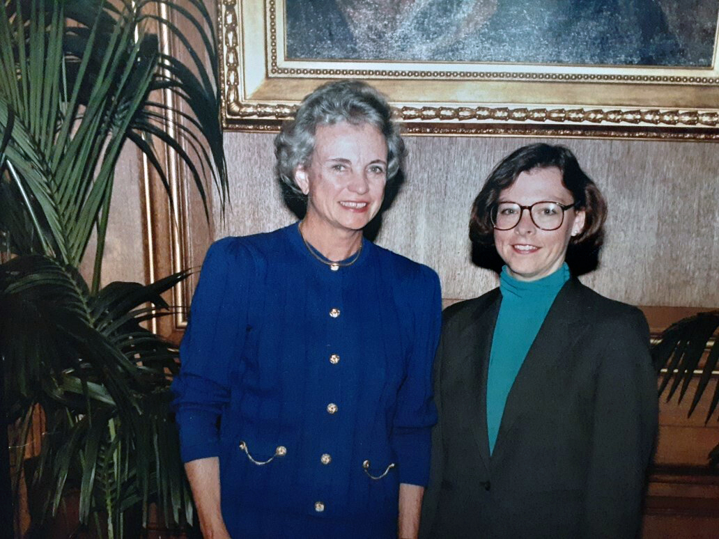 A woman in a blue dress with grey hair stands next to a woman in a black blazer and green mock turtle neck, with a potted palm on the right side of the woman in blue, and the bottom of a large gold picture frame behind them