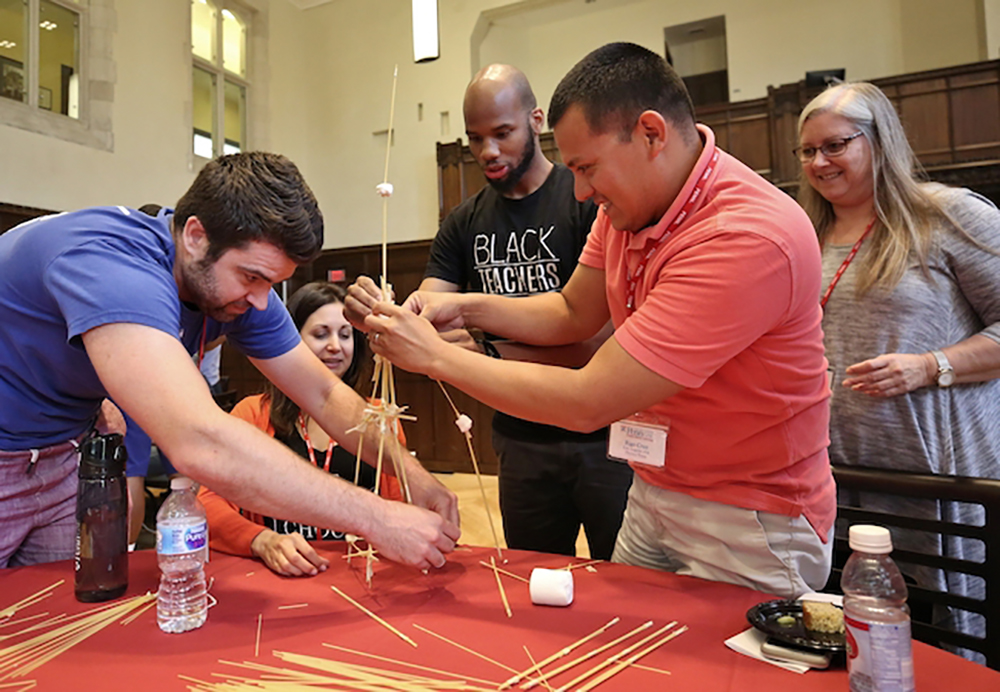 A group of educators in training work on a project building a tower of wooden sticks on a table.