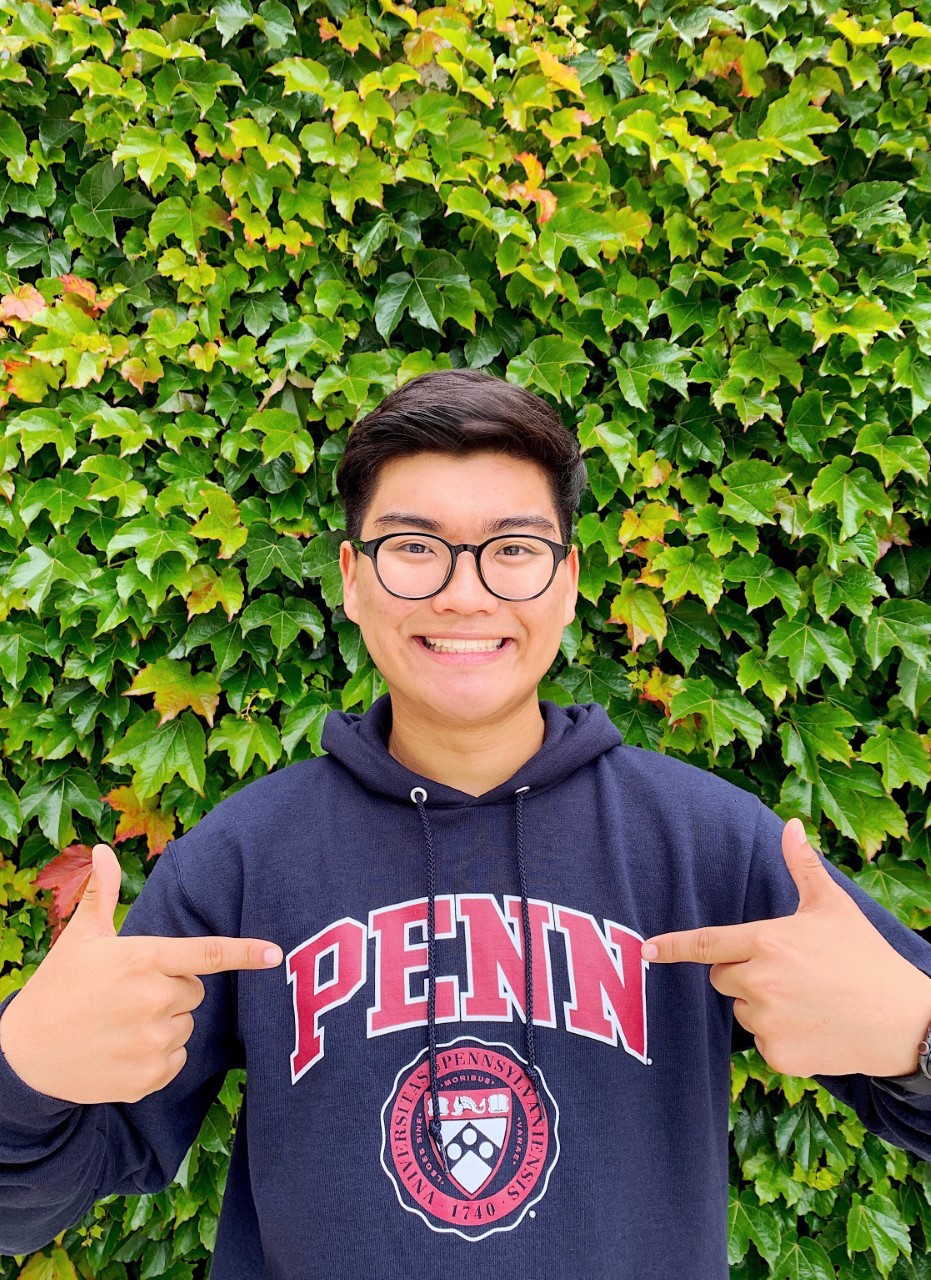 Man wearing glasses stands in front of an Ivy covered wall,  doing a thumbs up pose and smiling at the camera, wearing a blue sweatshirt with red lettering reading "Penn"