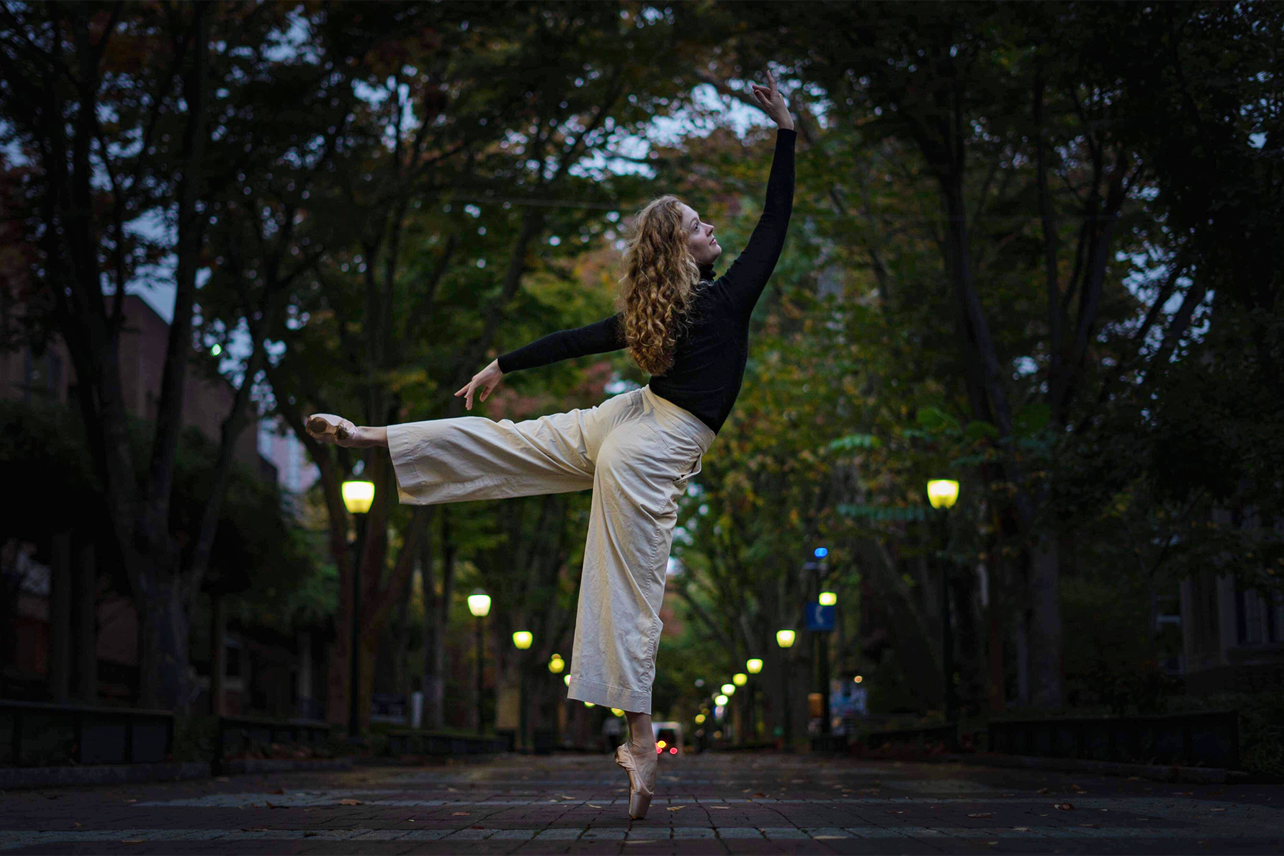 student on Locust Walk wearing ballet shoes and posing in a ballet position on her toes