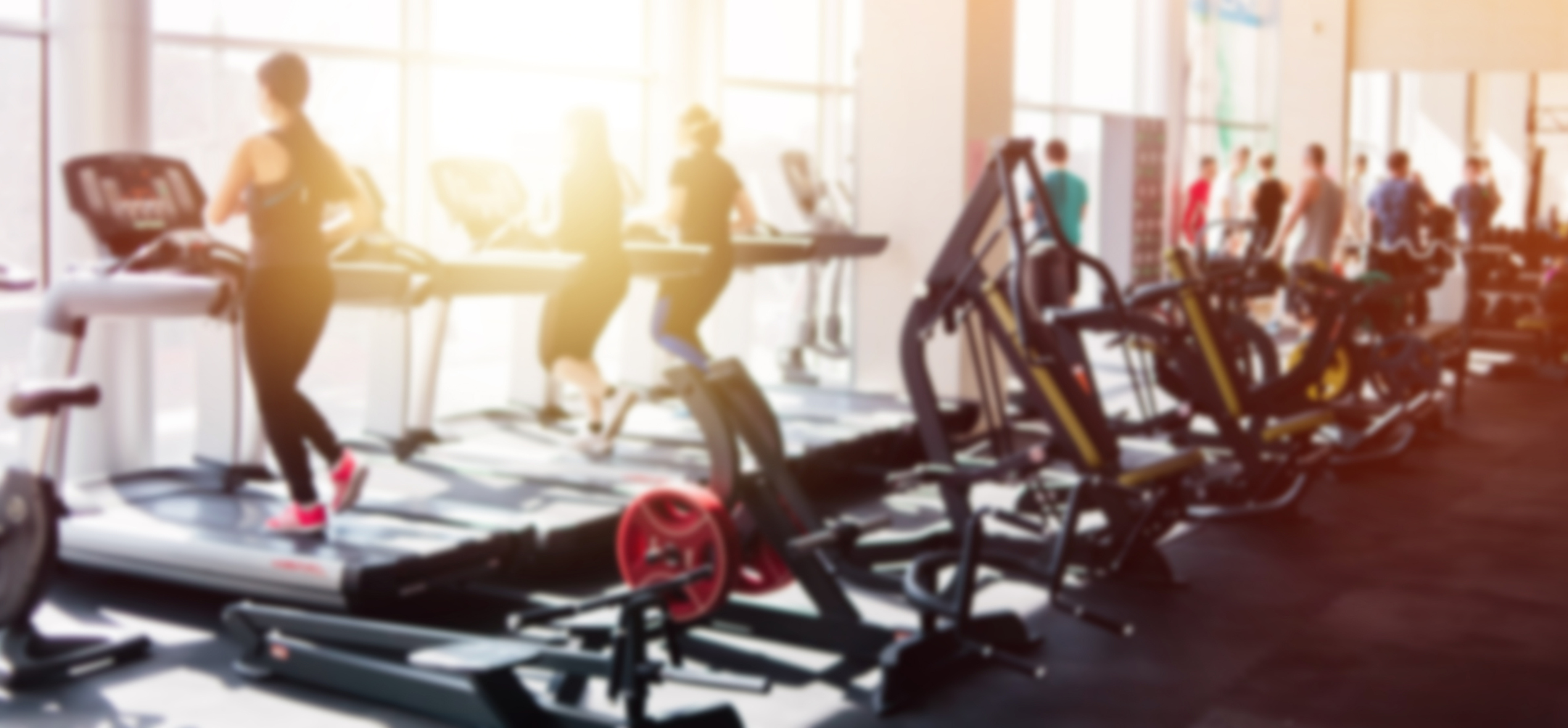 eople run on treadmills in a fitness center, with sunlight shining through a large glass window.