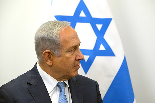 A man with short gray hair wearing a dark blue suit jacket, white shirt and light blue tie is seen in profile in front of an Israeli flag