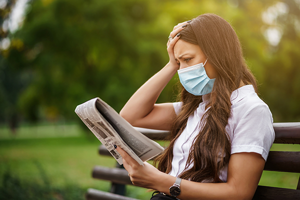 Person wearing a face mask seated on a park bench reads the newspaper while holding head in concern.