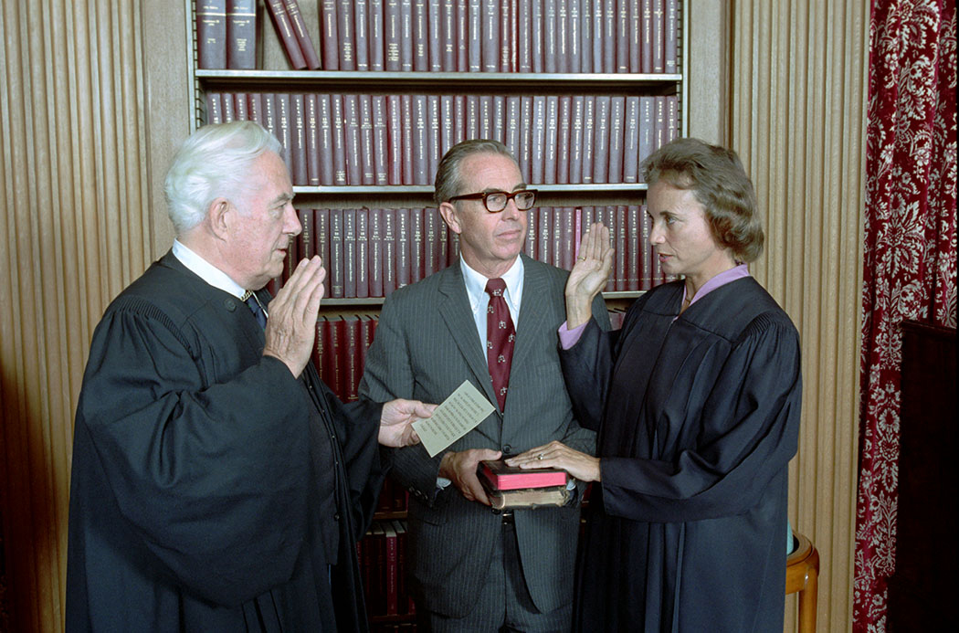 Three people stand in front of a bookcase full of books in burgundy binding, the man on the left is wearing judge robes and has his right hand in the air, the woman on the right is in judge robes and has her right hand in the air and left hand on a bible and a man in the middle wears a suit and tie, is holding the bible and is looking at the woman