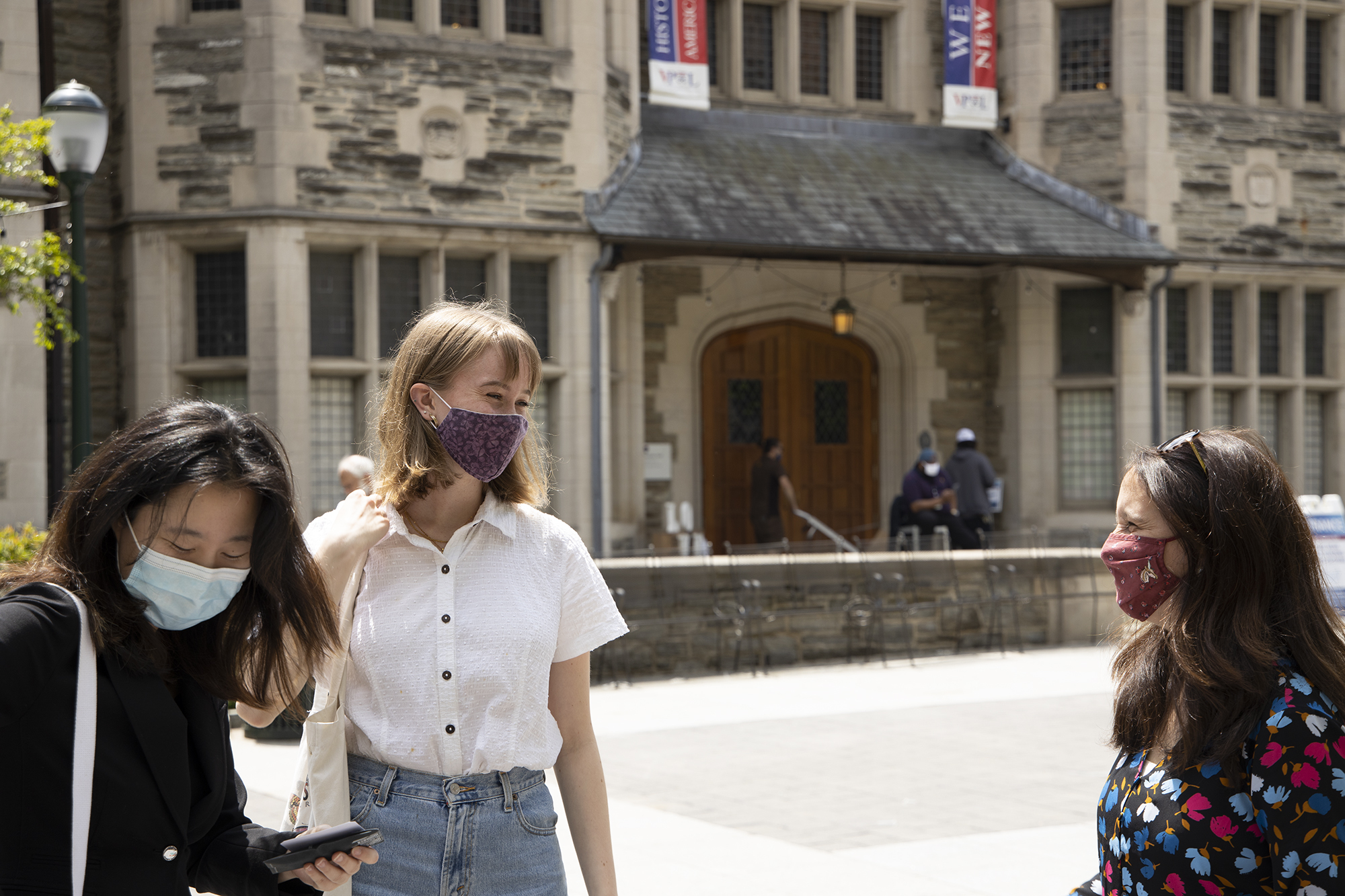 three women wearing masks smiles at each other in front of an old stone building