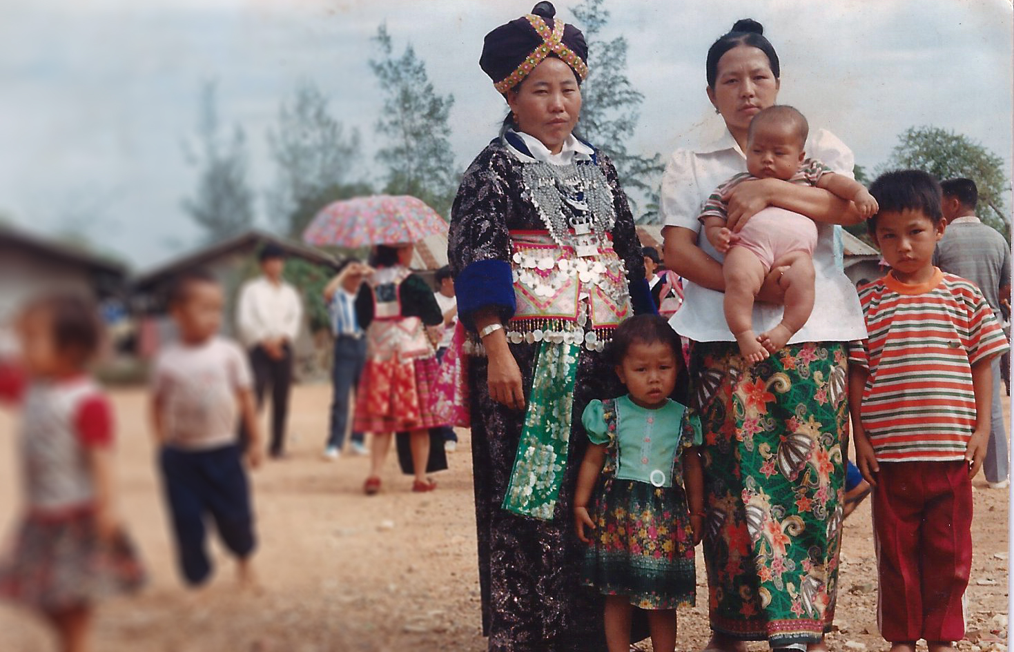 Two Hmong women in traditional clothing pose with two children and a baby. Trees, buildings, and people are seen in the background
