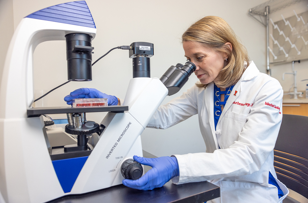 Sharlene M. Day looking in a microscope in a lab wearing a lab coat and latex gloves.