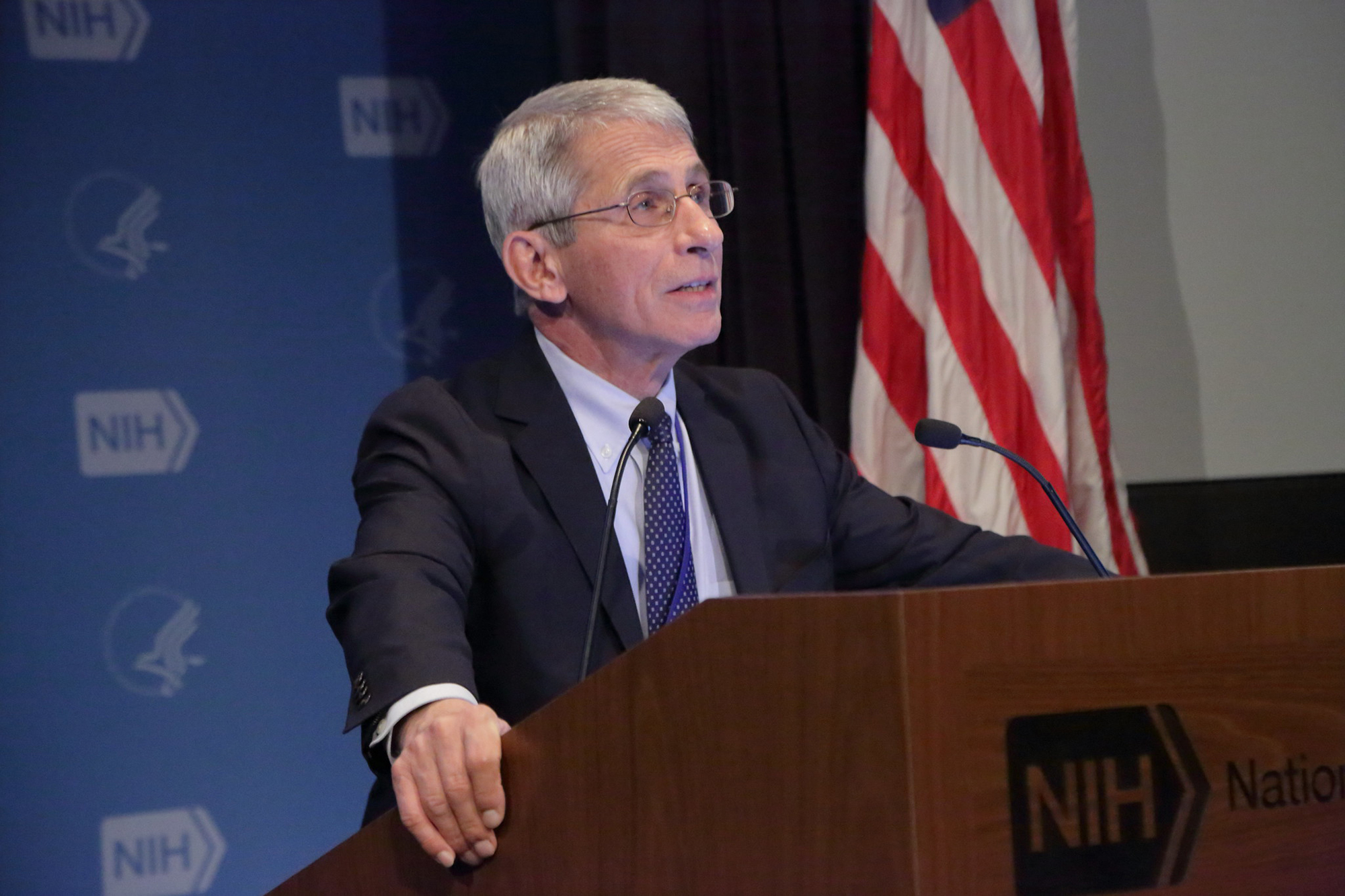 Anthony Fauci speaking at a podium with an American flag in the background.