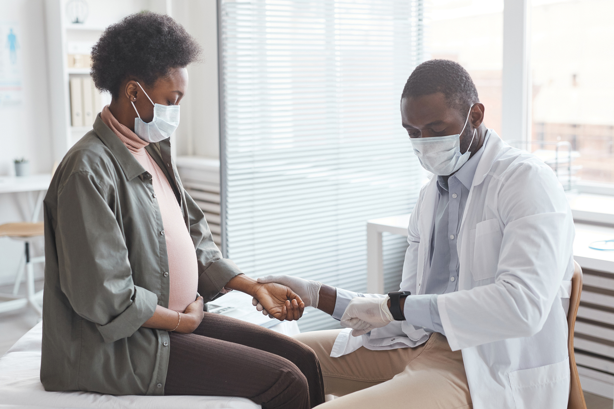 Pregnant person in a mask having their pulse checked by a masked doctor.