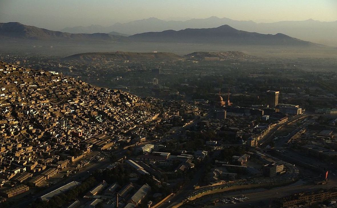 A city in the desert is seen from above, with brown structures dotting the valley and mountain ranges in the distance, some covered in haze.