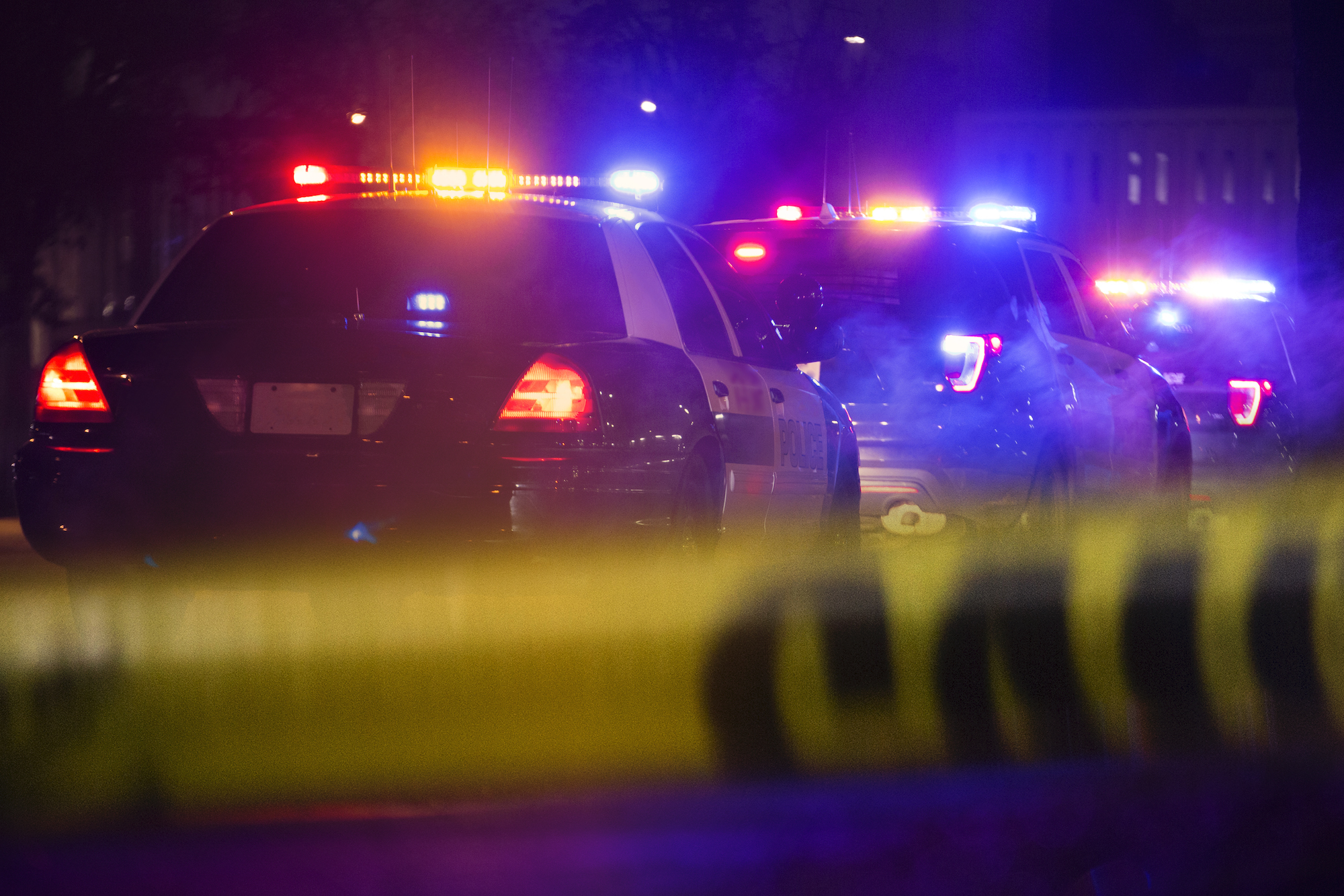 Three police cars with sirens flashing are seen in a line from behind police crime tape at night