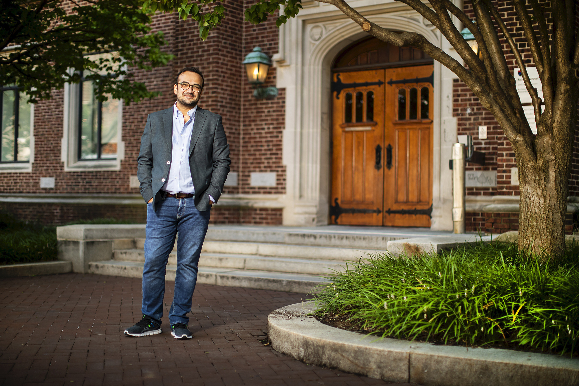 Hasan Küçük stands with his hands in his jeans pockets in front of the wooden double doors and red brick facade of  Fisher-Bennett Hall