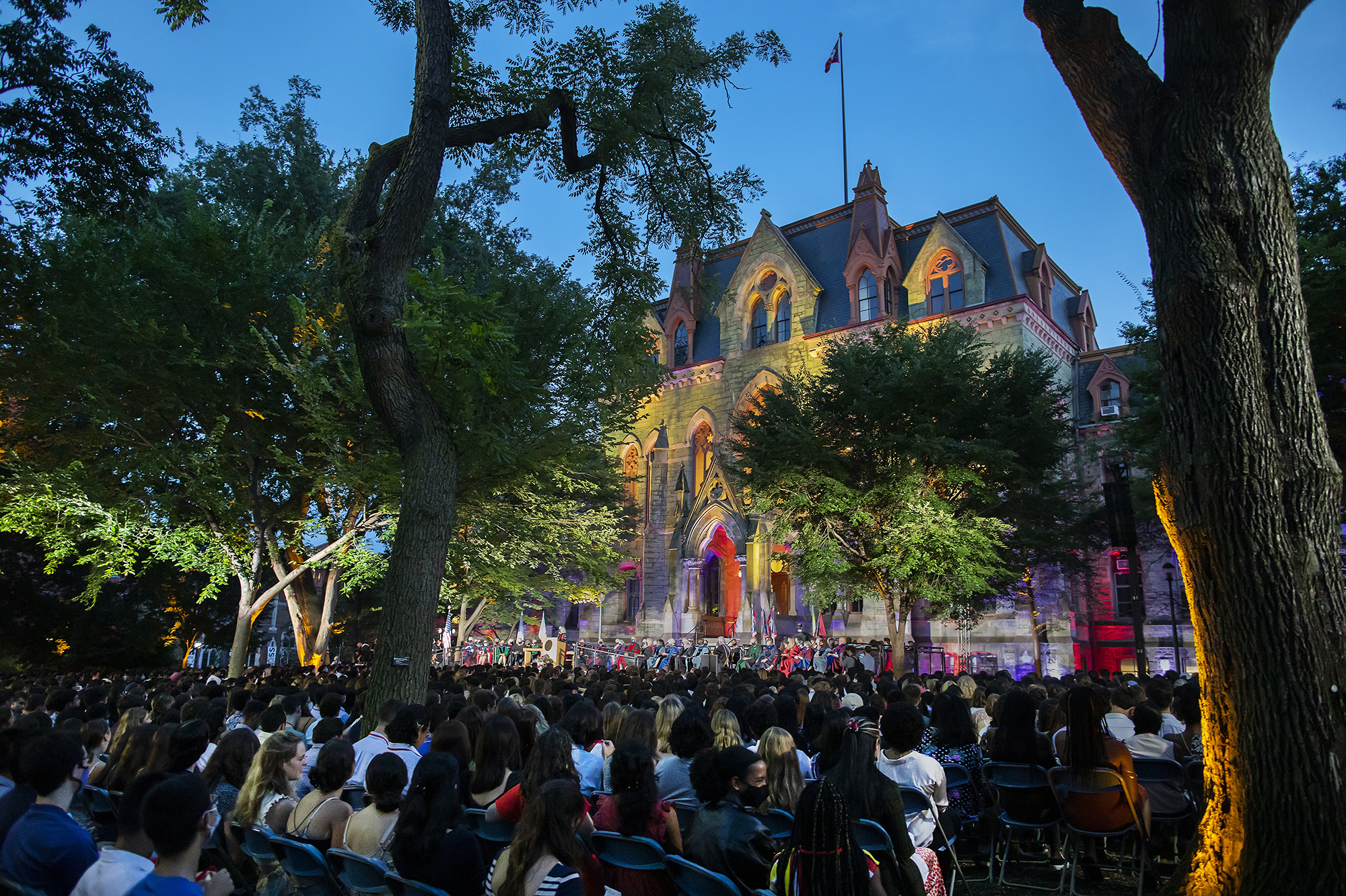 Crowd of students seated in front of College Hall lit with blue and red lights at dusk.