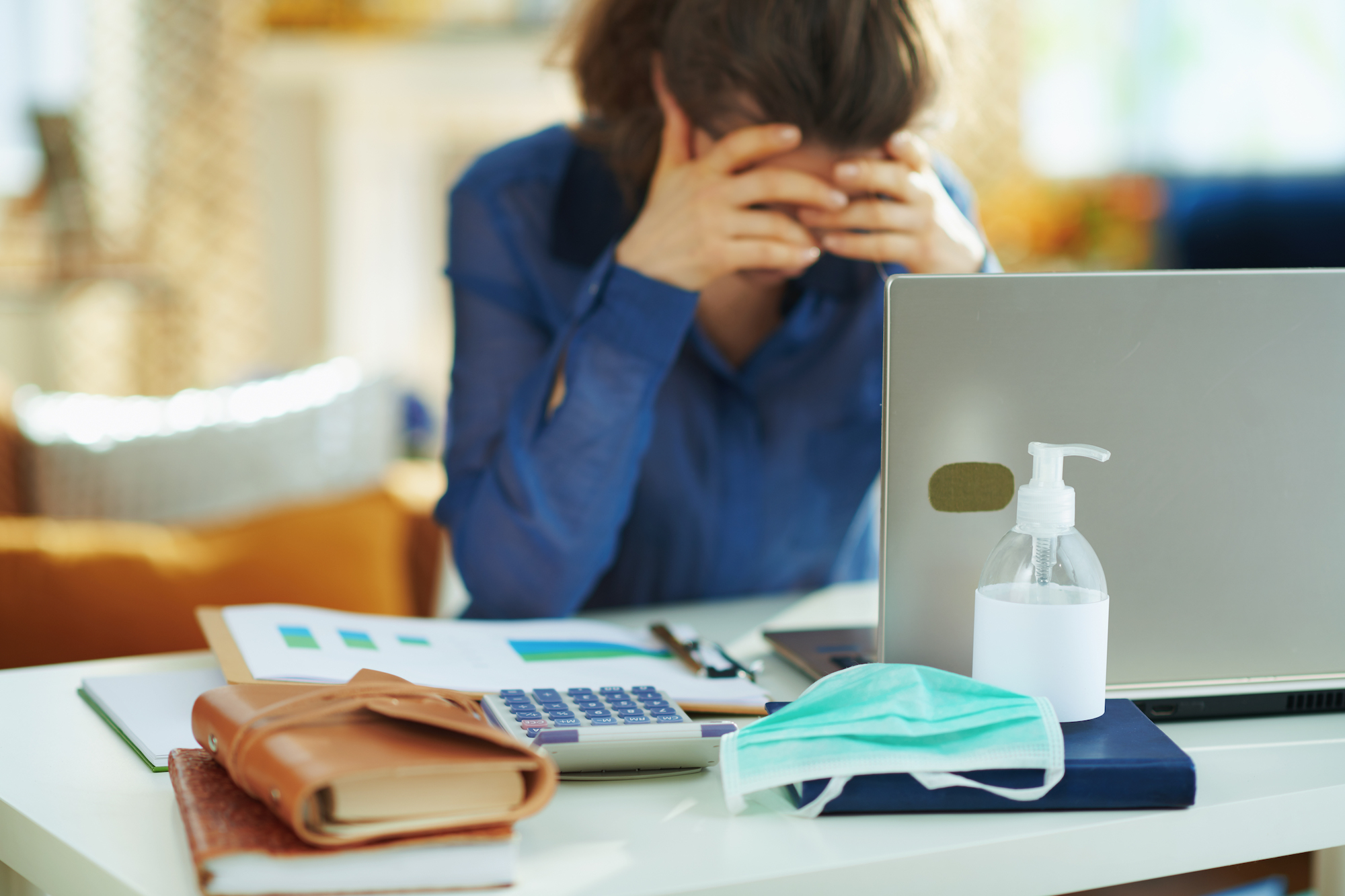 Person holds head in their hands sitting behind a laptop screen with hand sanitizer, a face mask, a calculator and a wallet in the front of the laptop.