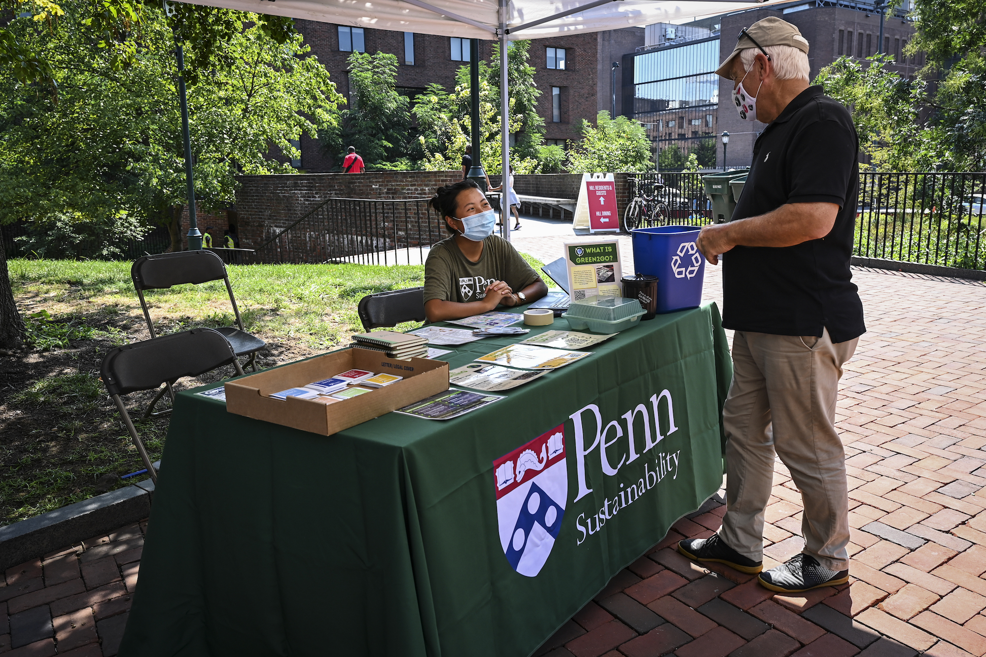 A person seated at a table labeled Penn Sustainability interacts with someone visiting the table