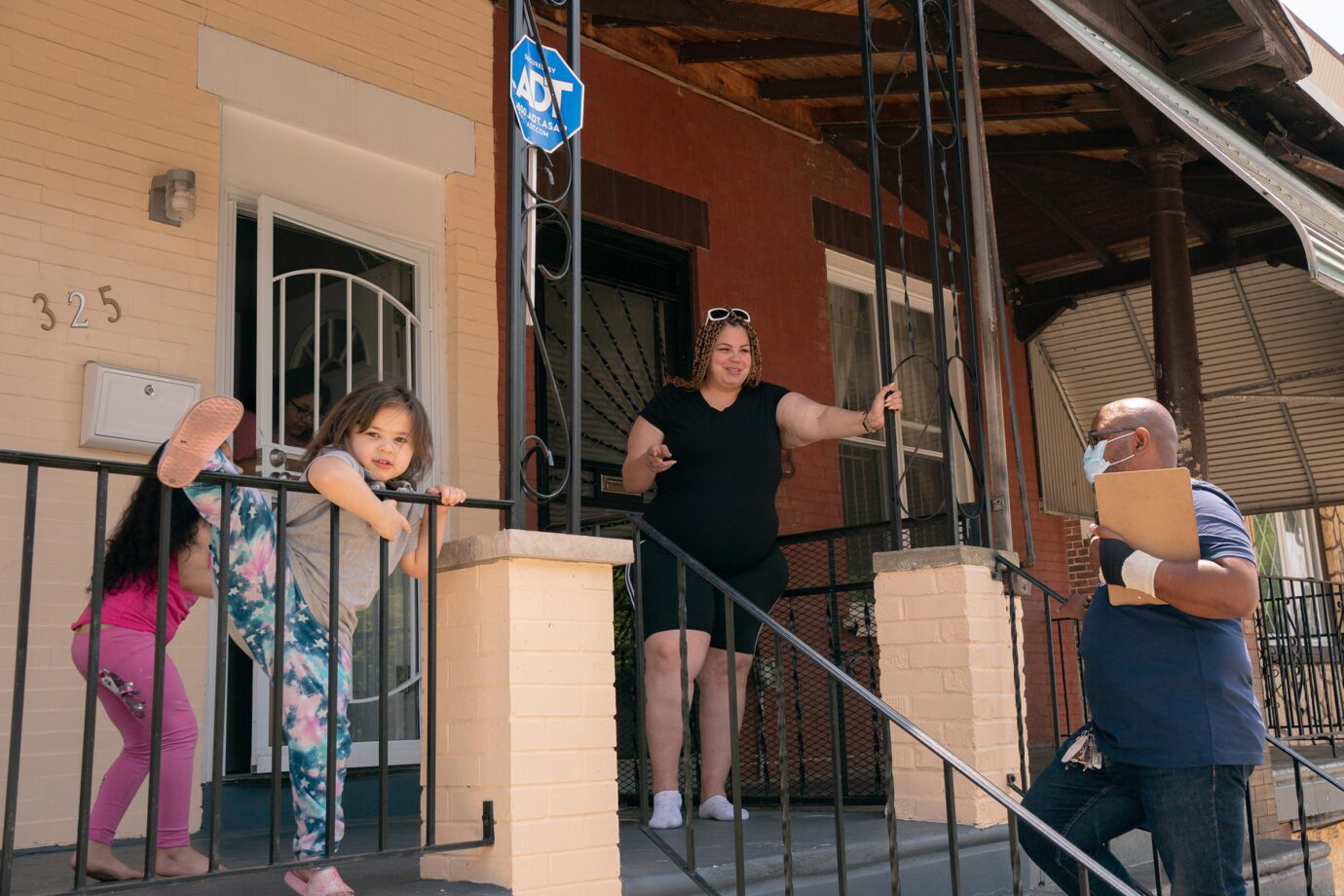 A person in a mask holding a clipboard at the bottom of steps outside a home. On the porch are an unmasked adult and two unmasked children.