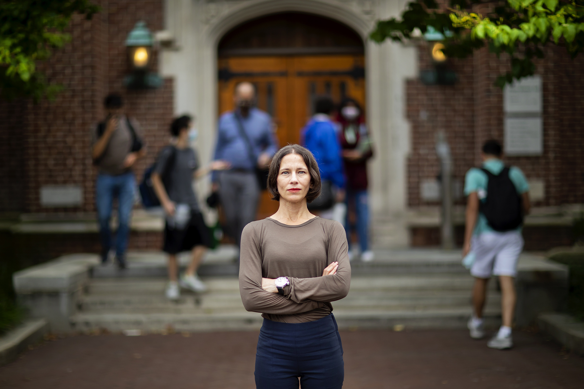 A woman stands with folded arms in front of a building; six people enter/exit at the doors behind her