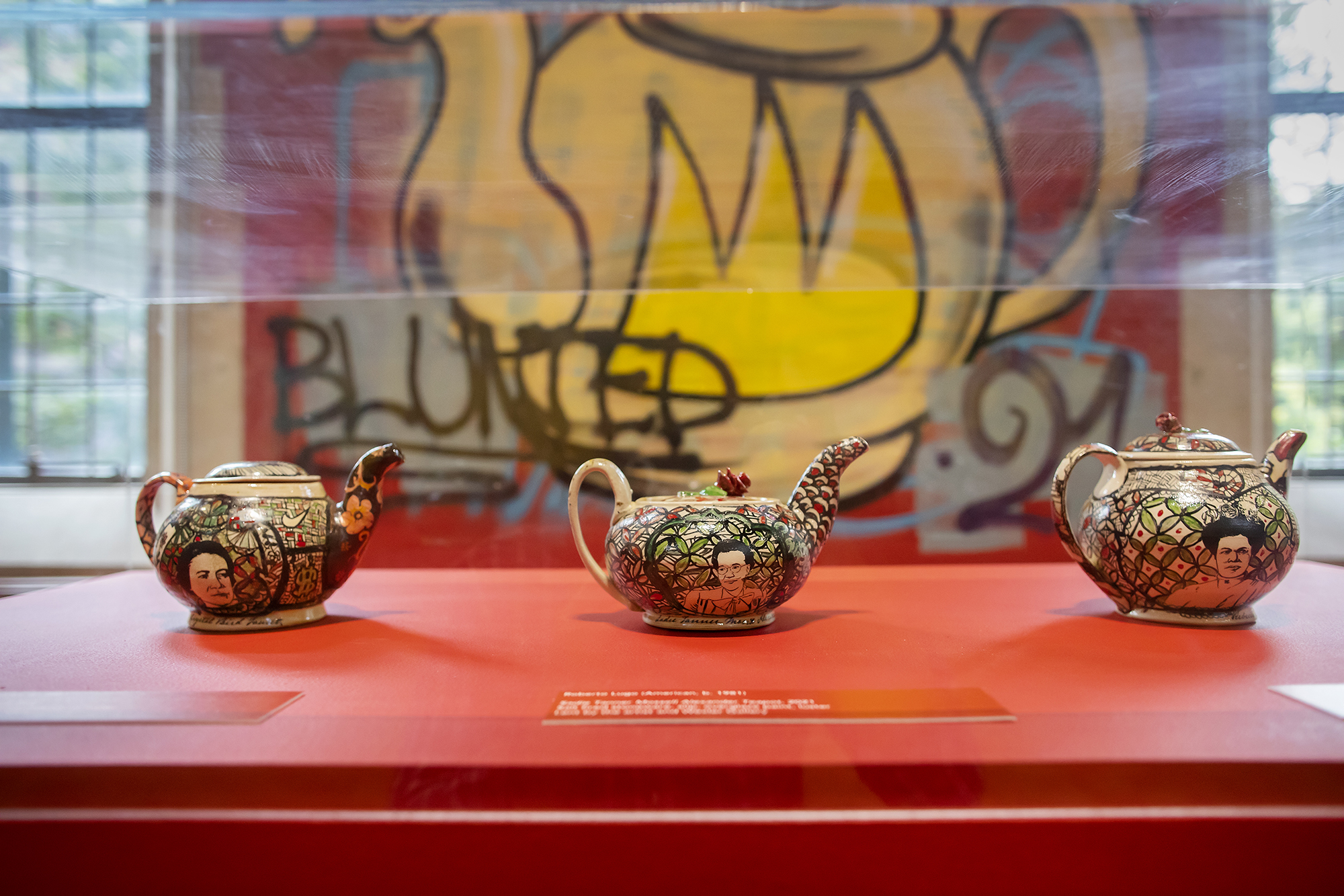 three ceramic teapots in a case with graffiti of a pot behind them 