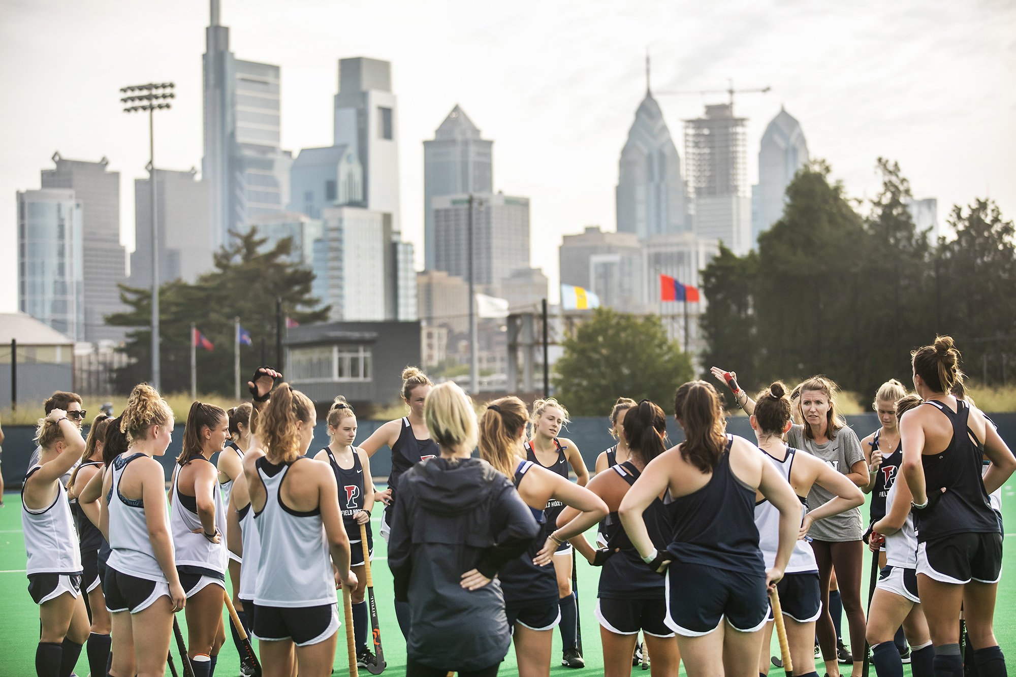 With the Philadelphia skyline in the background, Coach Colleen Fink addresses her players in a circle at Vagelos Field. All are standing.