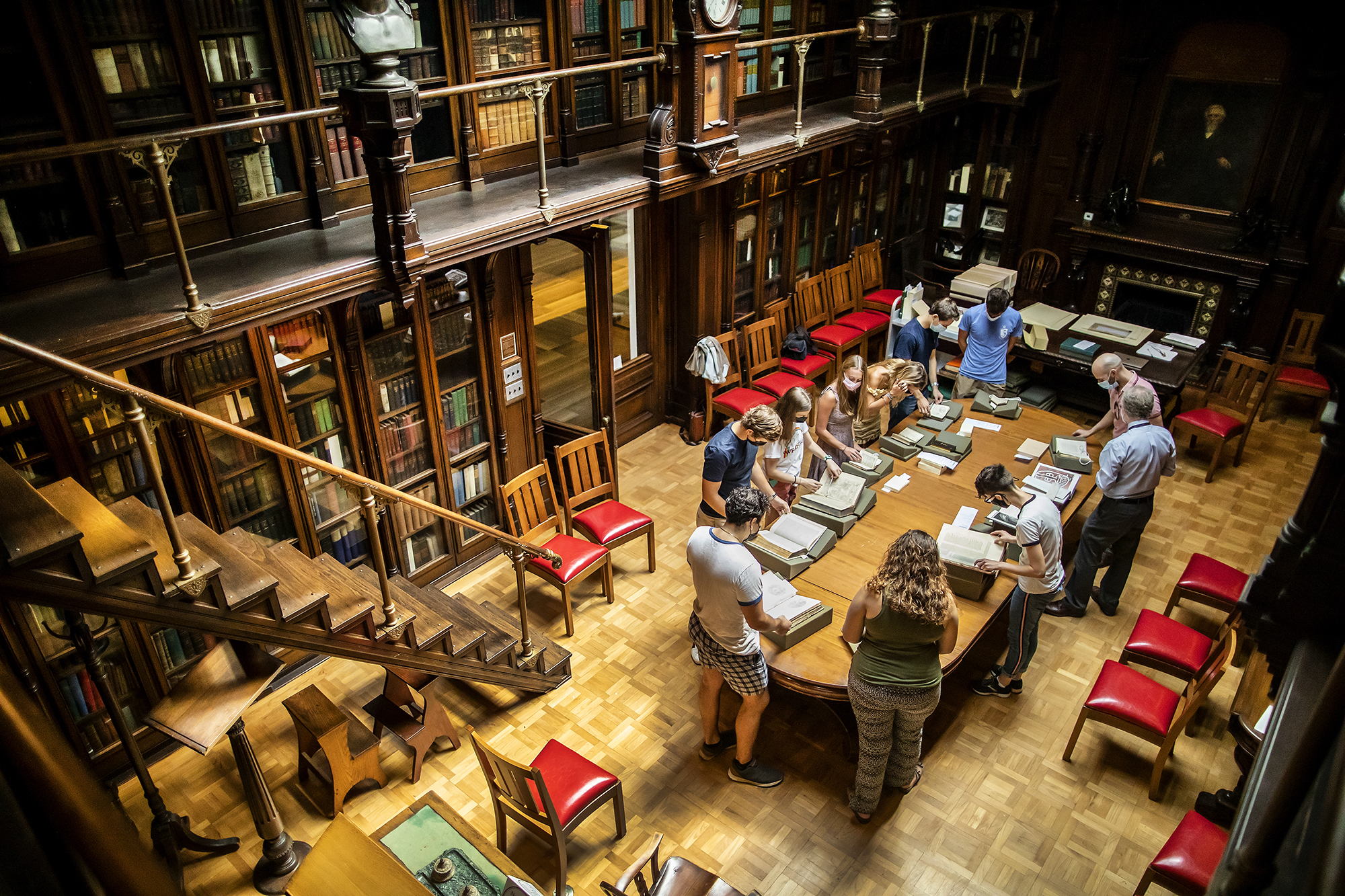 A room shot of class looking at books in a library with wooden floor, trim, and bookcases