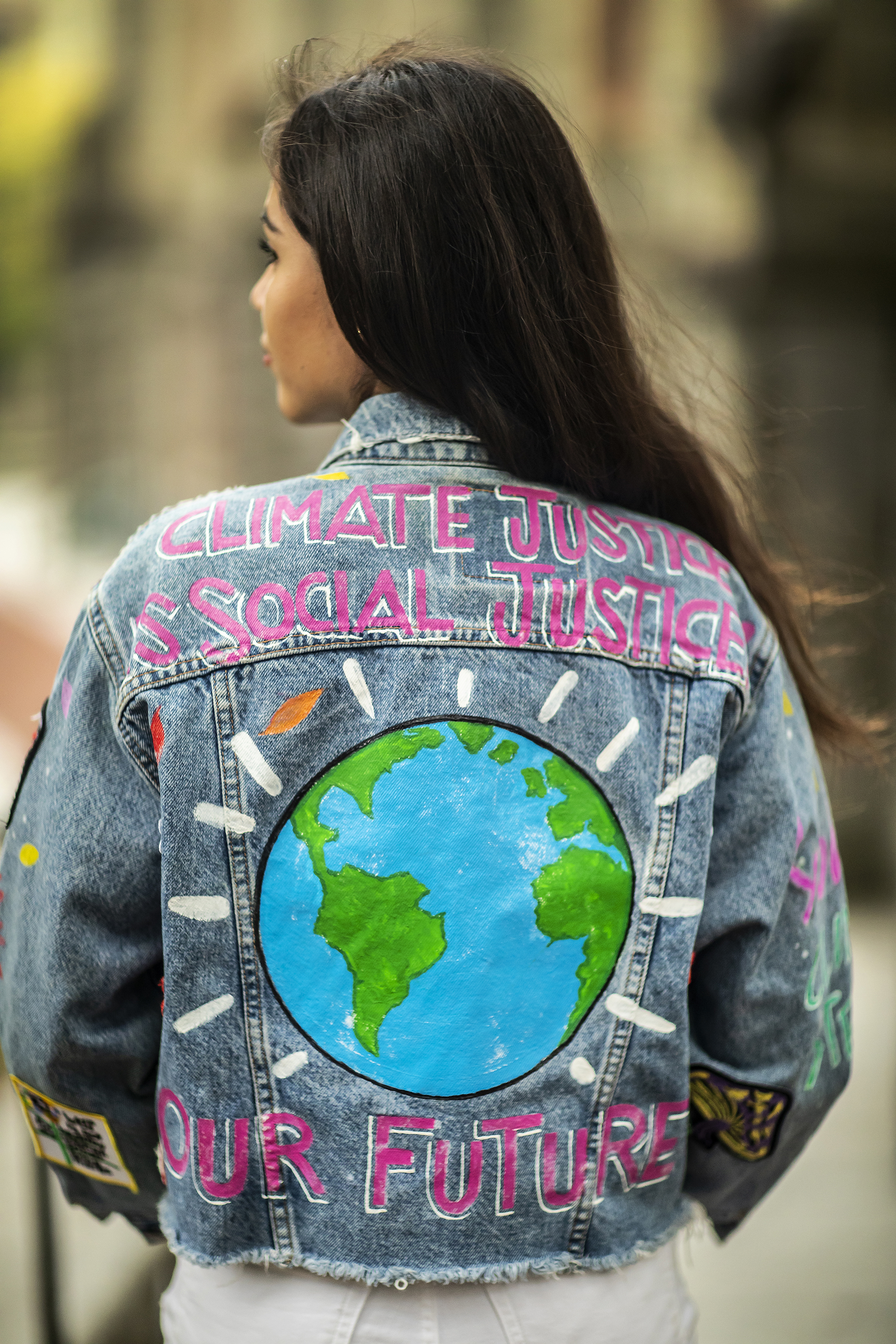The back of a college-age woman wearing a jean jacket decorated with a blue-and-green globe and the words "Climate Justice is Social Justice" and "Our Future."