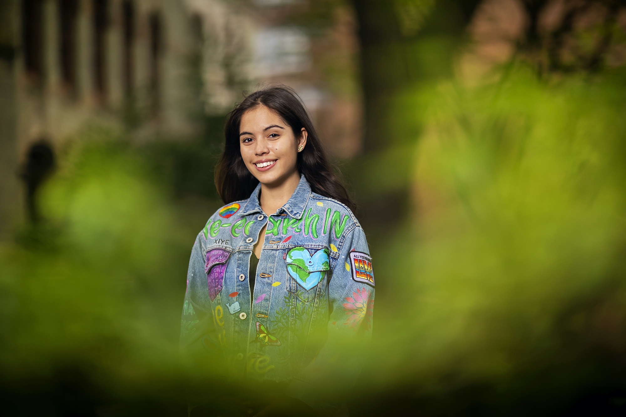 A college-age person standing outside, with greenery blurred in the front of the image. She is wearing a jean jacket with the words "Re-earth IN," a globe in the shape of a heart, and other earth-related designs. 
