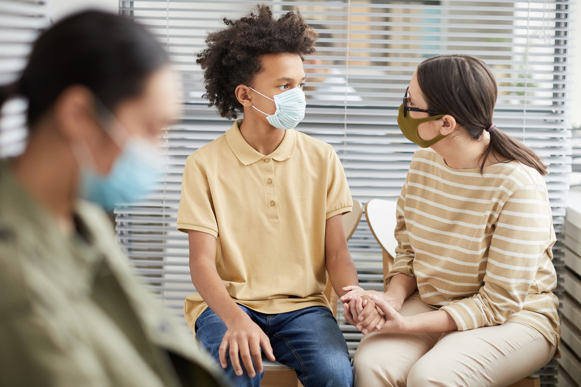 Adolescent wearing a mask seated in a waiting room with an adult in a mask holding their hand.