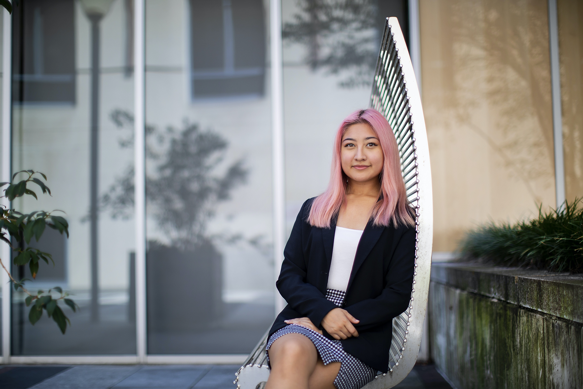 A person sitting outside on a silver metal bench wearing a black and white skirt, a white shirt, and blue blazer. Plants are visible to the right and to the left. 