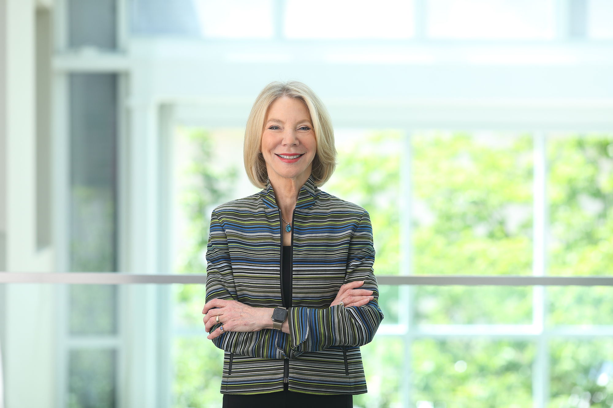 Amy Gutmann stands with arms crossed by a sunlit window.