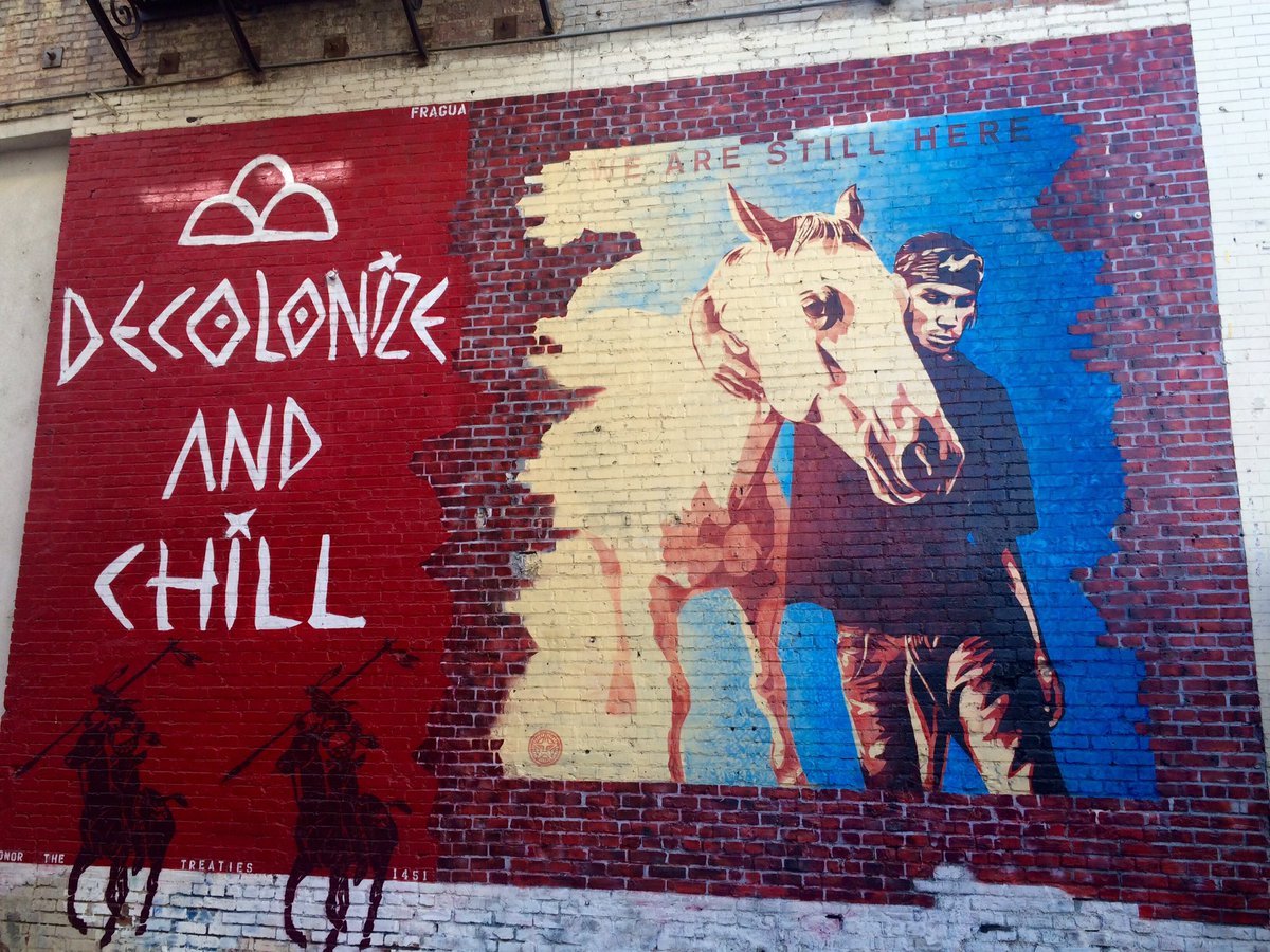 A mural on a brick wall. On the left-hand side, on a red background, are the word "Decolonize and Chill" in white. Beneath that are the silhouette of two men riding horses. On the right-hand side is a person with a horse, beneath the words "We are still here."