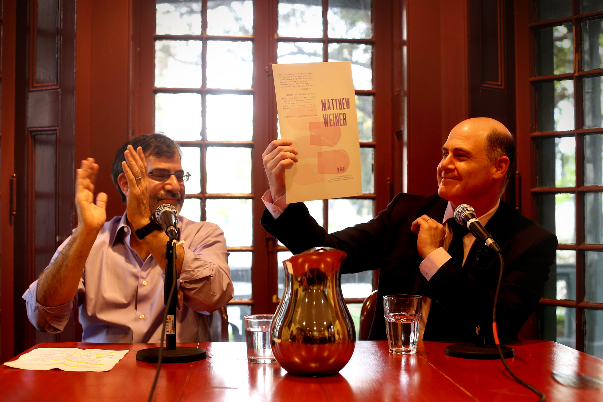 Al Filreis claps, seated next to Matthew Weiner, who is holding up a flyer, at the Kelly Writers House.