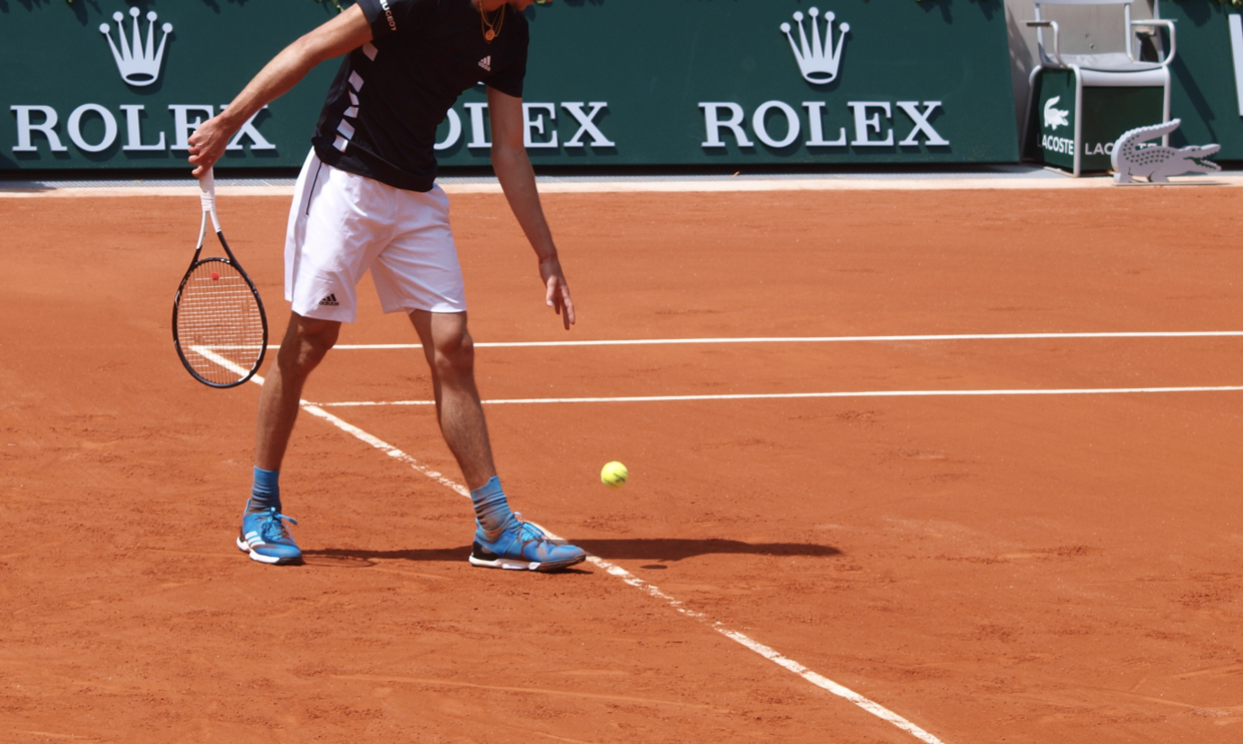 Tennis player preparing to serve on a clay court with Rolex signs on wall of court in background.