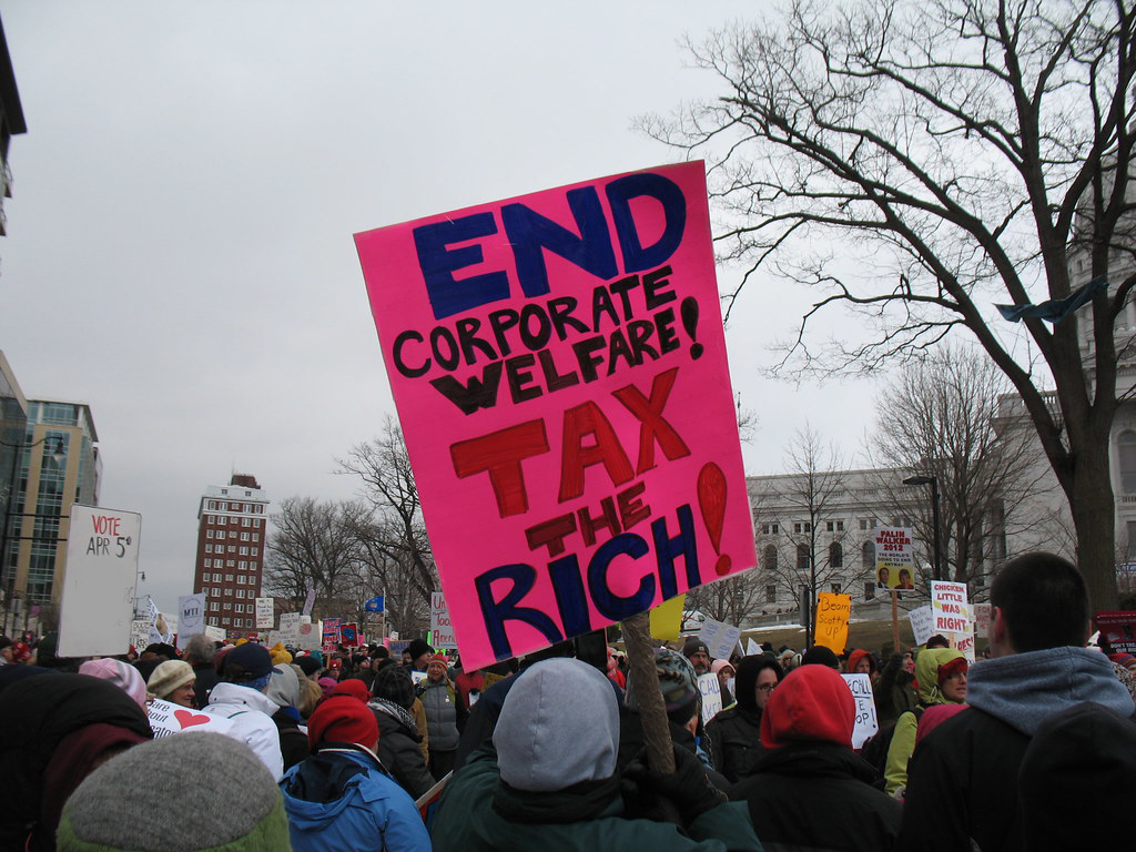protesters are seen from behind on a city street, one holding a hot pink sign with handlettering reading "end corporate welfare! tax the rich!