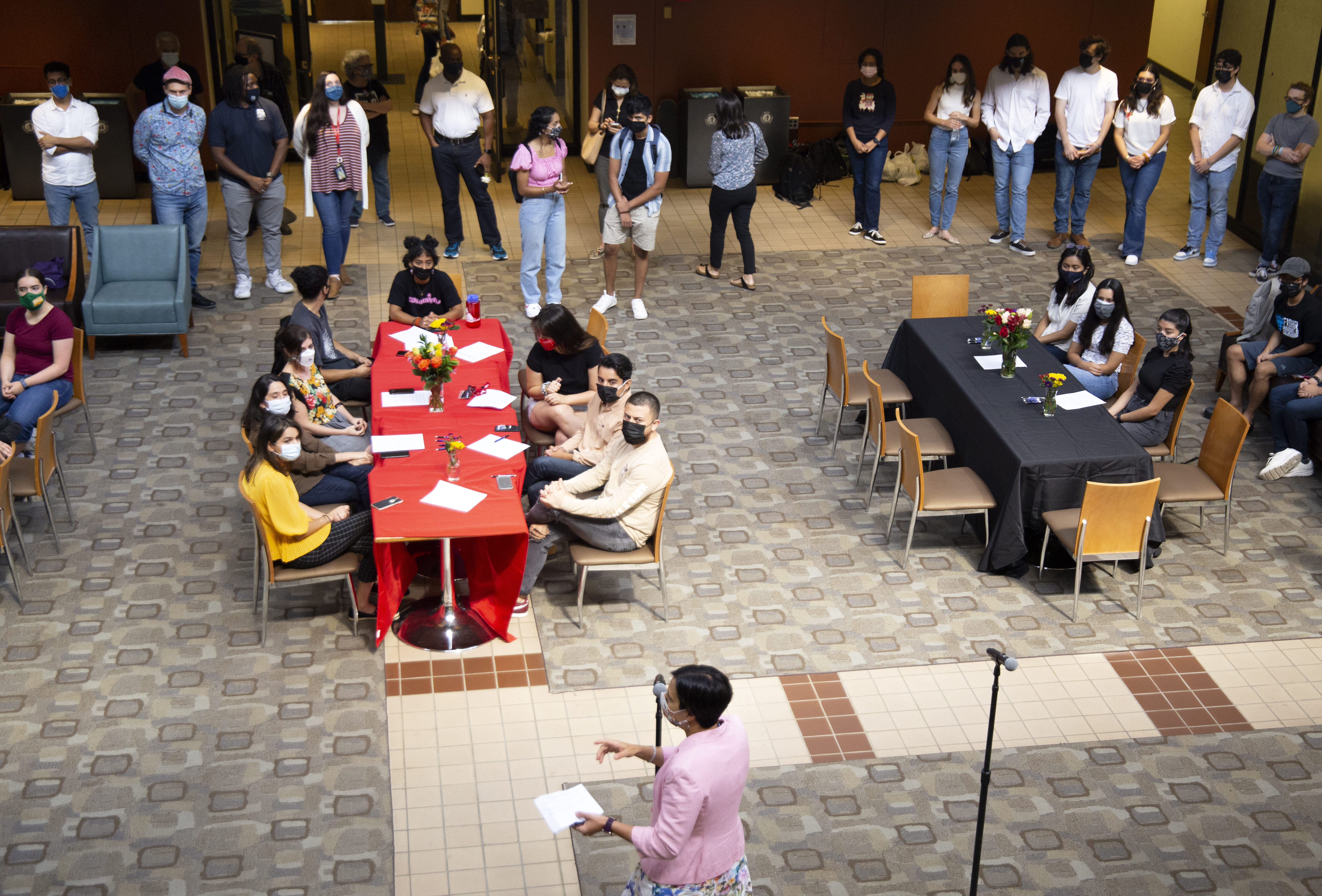 A woman in front of a microphone speaks to groups of seated and standing students