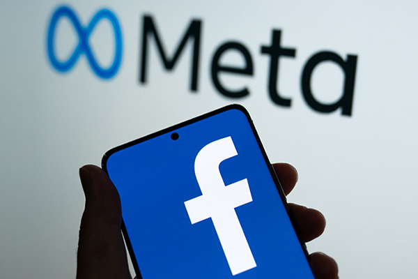 Hand holding a smartphone with the Facebook logo, in the background is the new Meta logo.