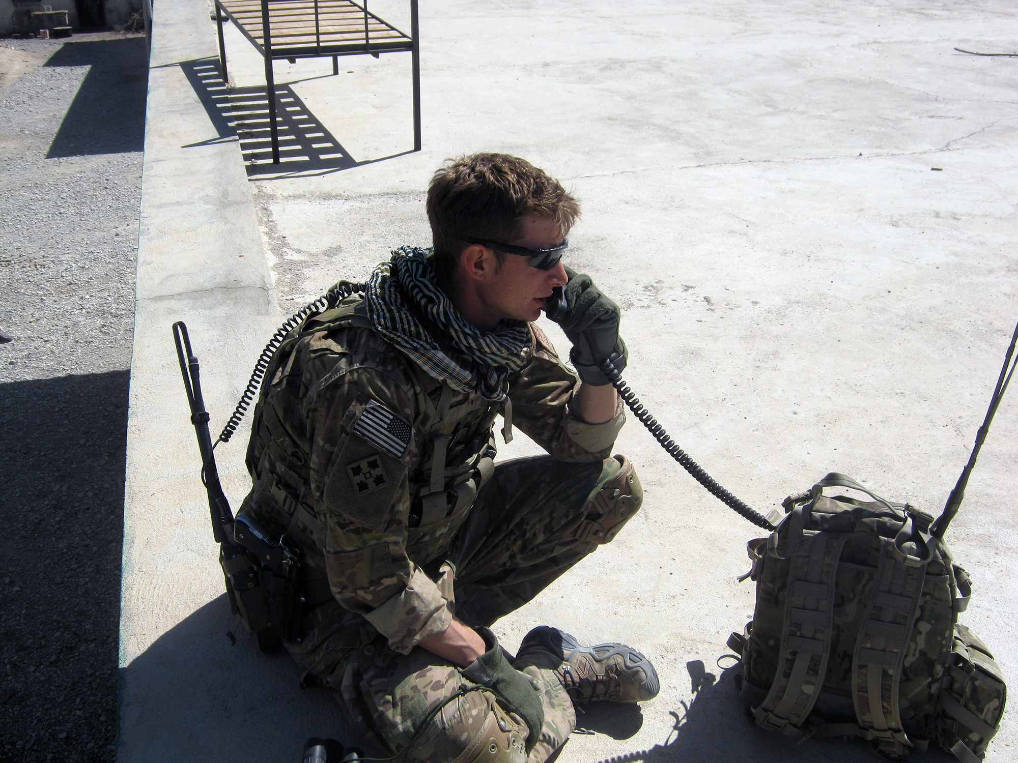 A person in combat fatigues sits on the ground speaking on a sat phone.