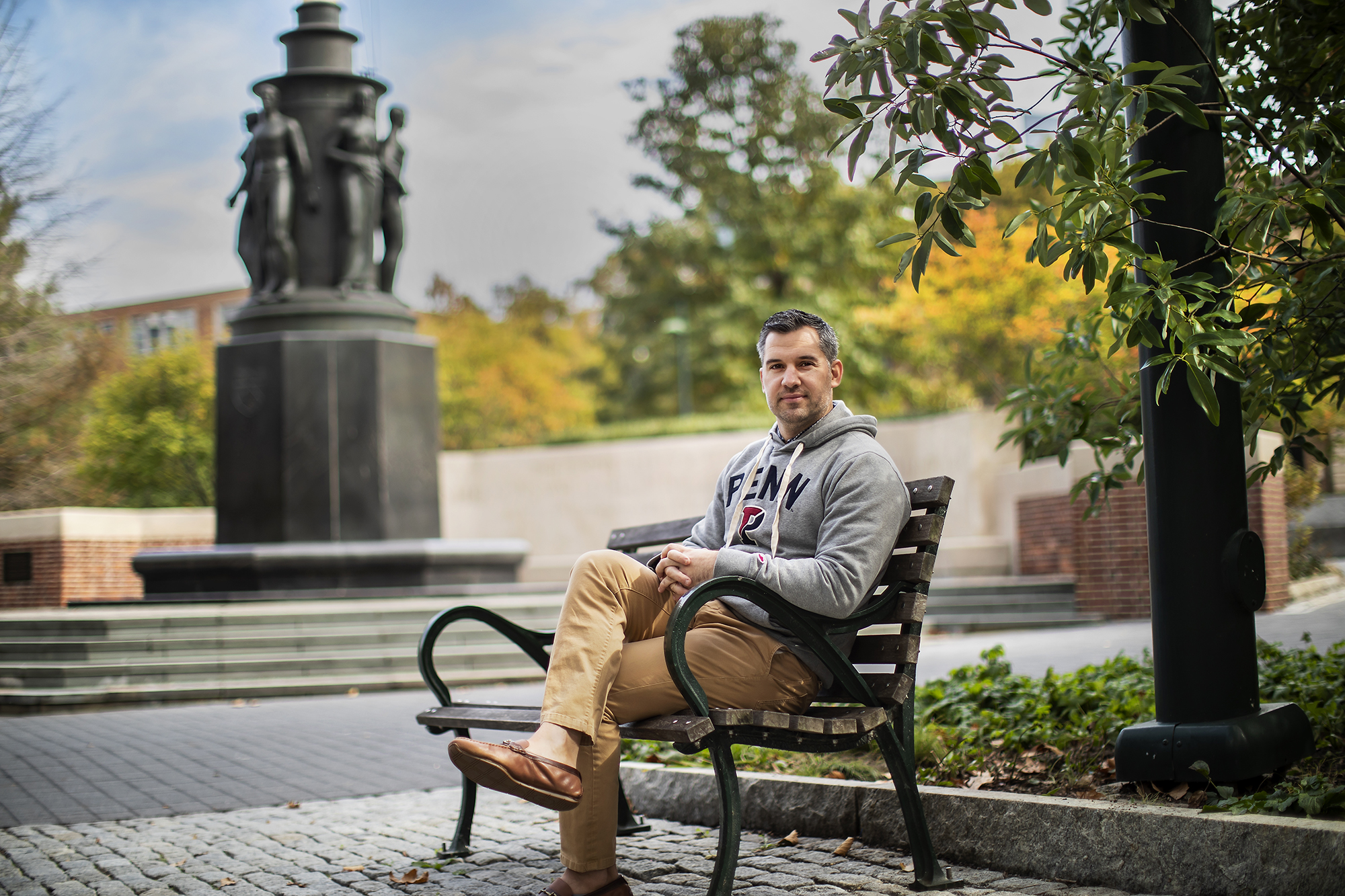 Greg Lutmann sits on a bench in on campus near a statue.
