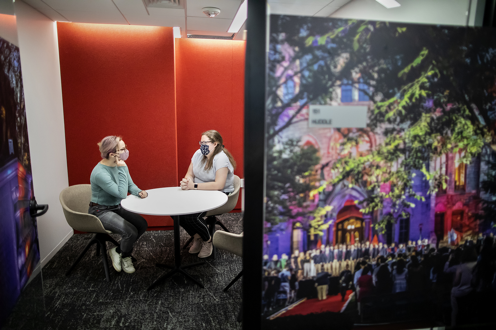 Two women wearing masks sit at a white round table in front of a red wall, with a colorful photograph of Penn's College Hall in the foreground