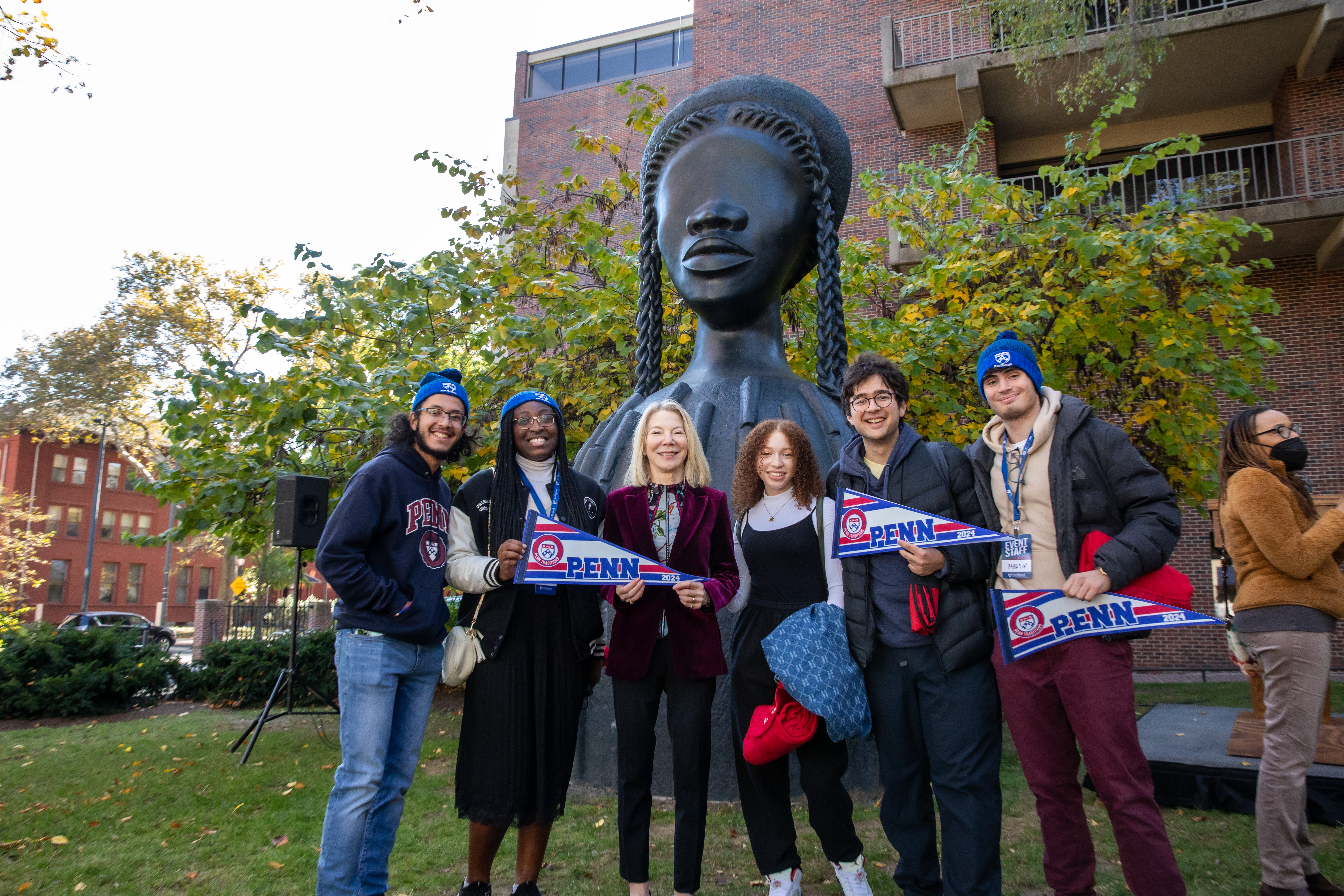 Gutmann and students smile for a photo in front of sculpture, holding logoed Penn flags with "2024" on them